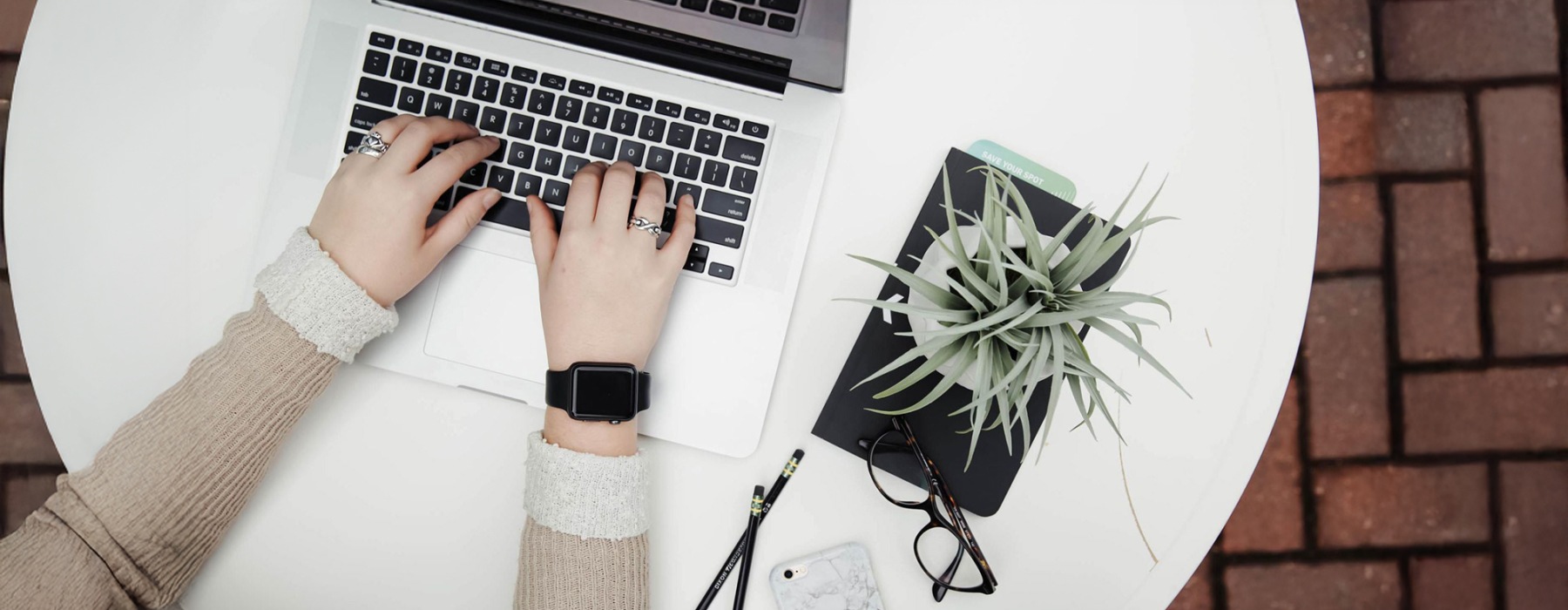 overhead shot of woman working on a laptop at a round table with pencils, her cell phone, glasses, notebook and a potted plant